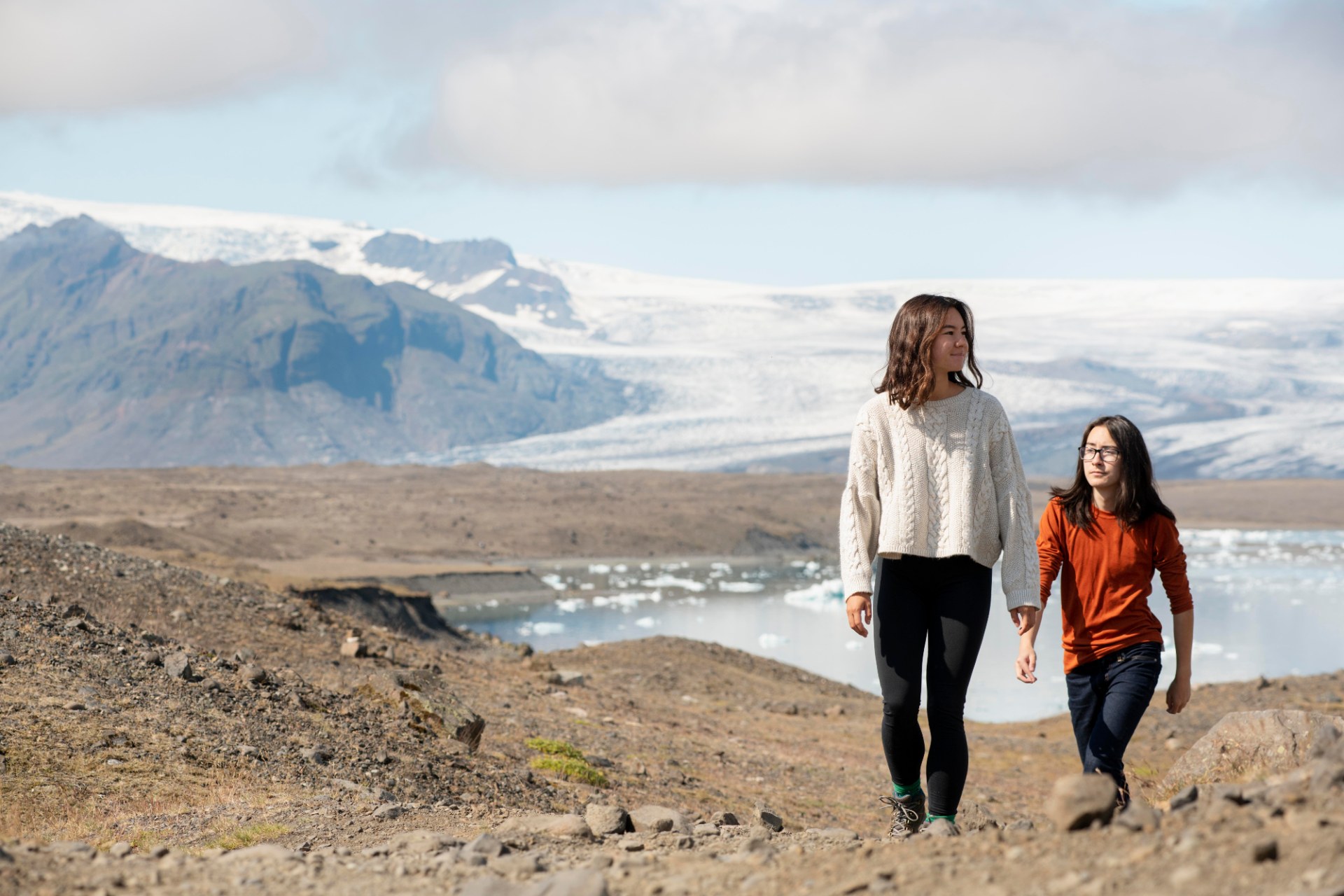 Duas jovens a fazer uma caminhada na Islândia, com um glaciar e montanhas ao fundo