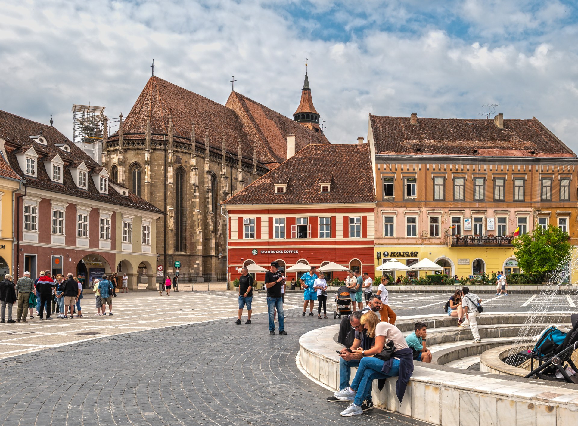 Jovens a descontrair junto à fonte na Praça do Conselho, em Brașov, com a Igreja Negra em fundo