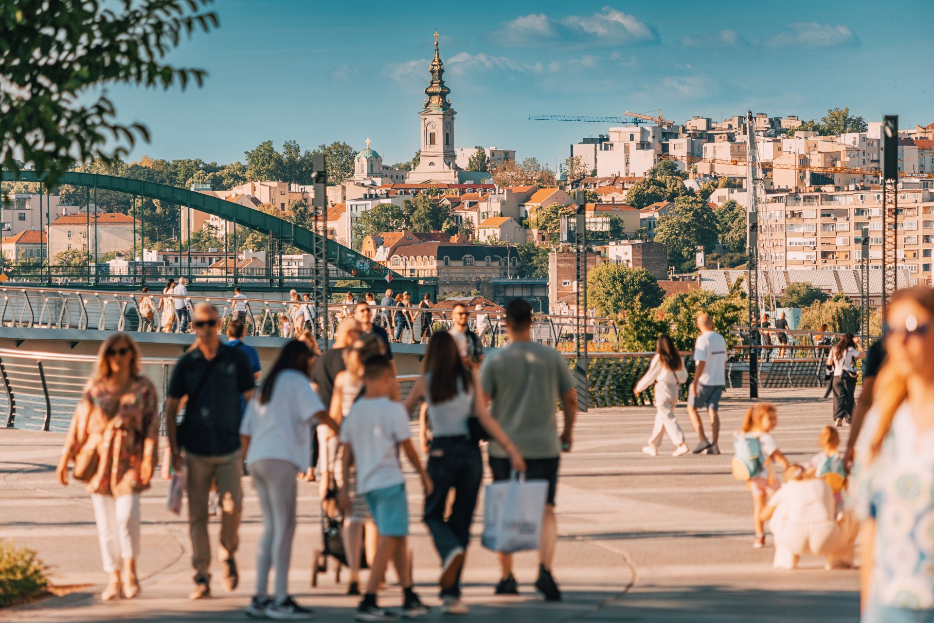 Pessoas a passear no passeio junto ao rio Sava, em Belgrado, com a cidade velha e a torre de uma igreja em fundo